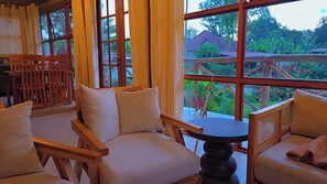 A room with a wooden sofa set, a round table, and large windows offering a view of a garden and buildings.