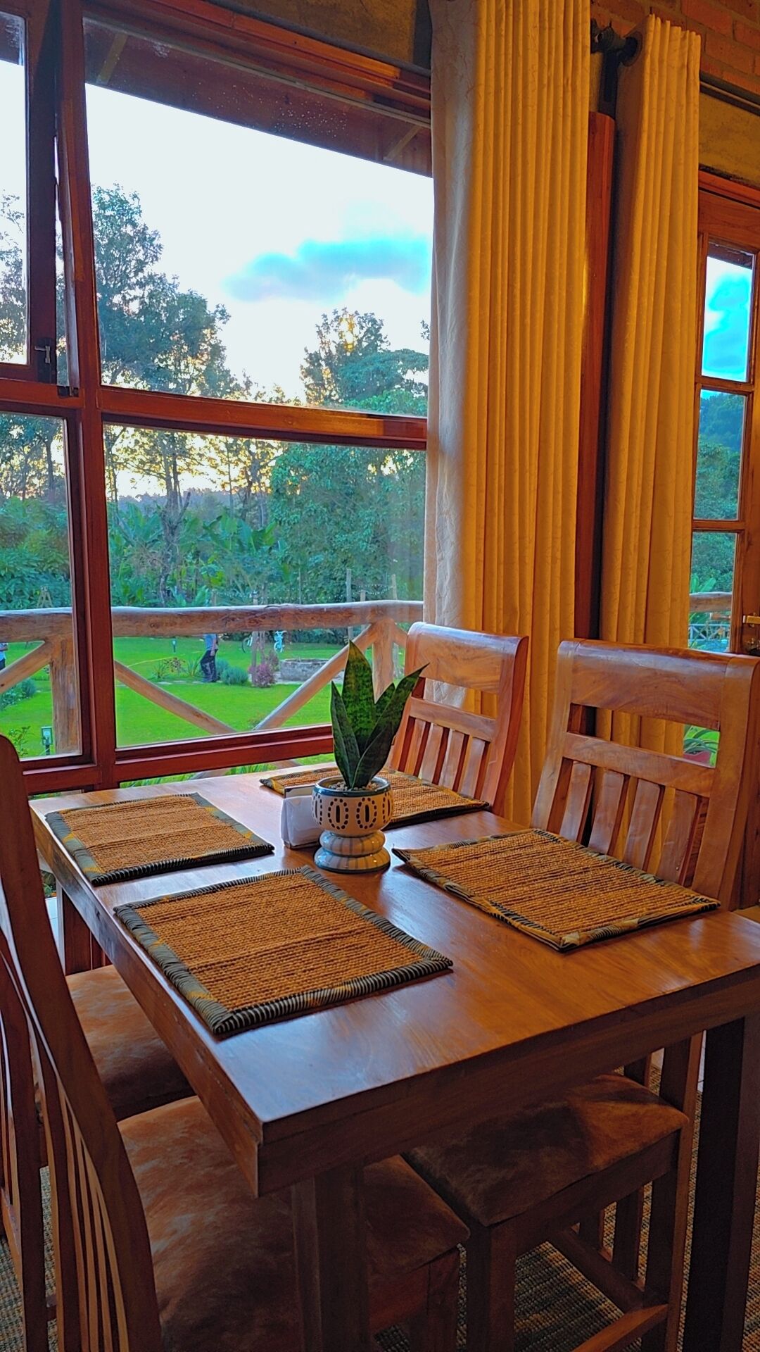 A wooden dining table with four chairs, a potted plant, and a view of a green landscape through large windows.