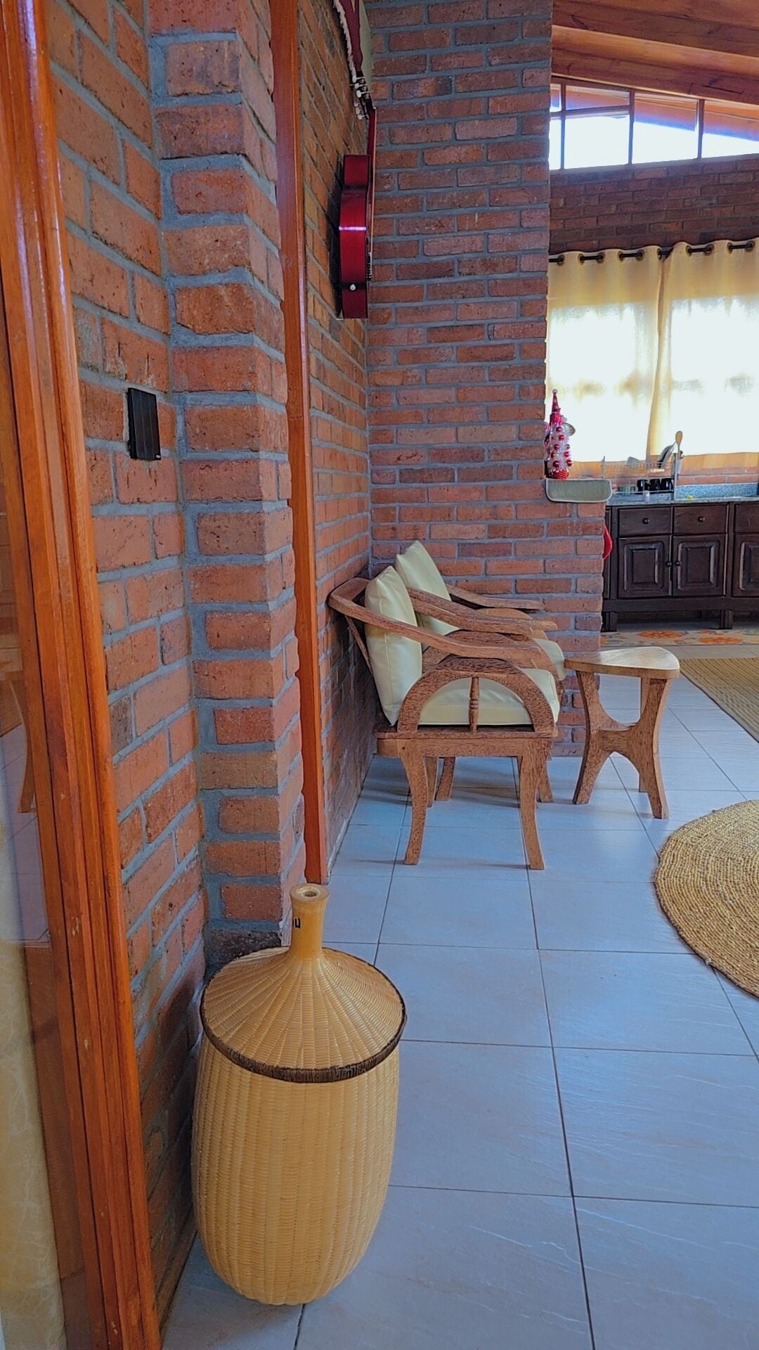 A corner of a room with a brick wall, a wooden bench with cushions, a woven basket, and a tiled floor.