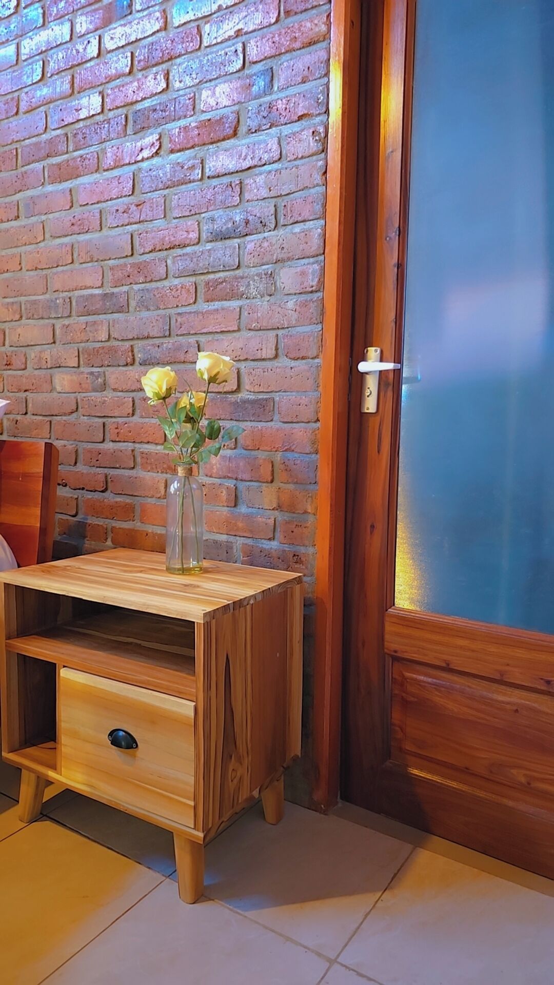 A wooden side table with a vase of yellow flowers, a wooden chair, and a brick wall.