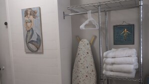 Un baño con una puerta blanca, un cuadro de una sirena, una tabla de planchar, un perchero con toallas dobladas y un cabezal de ducha colgante.