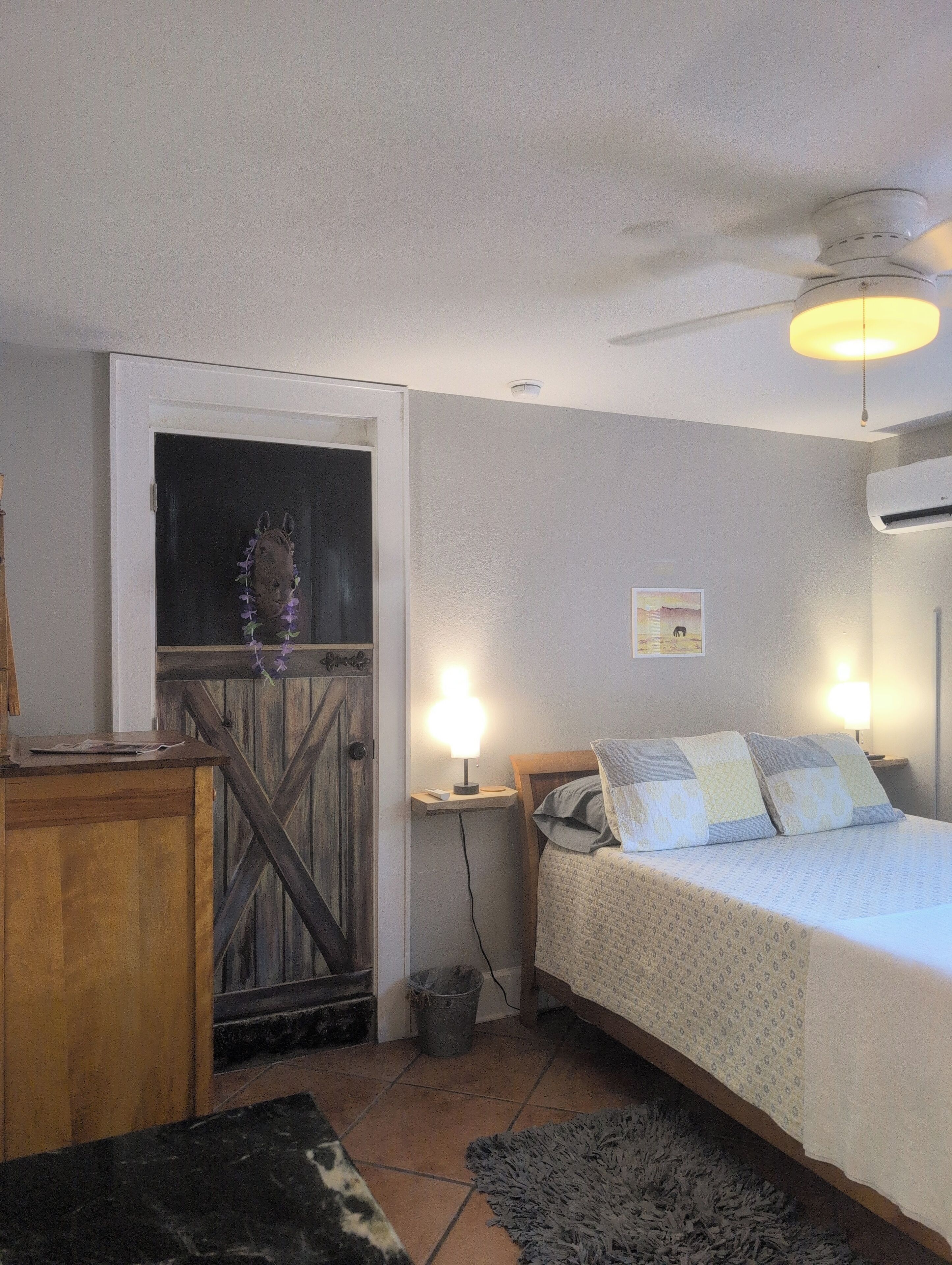 A bedroom with a bed, bedside lamps, a ceiling fan, a wooden door, and a small table.