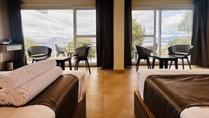A hotel room with two beds, a desk, two chairs, and large windows with a view of mountains.
