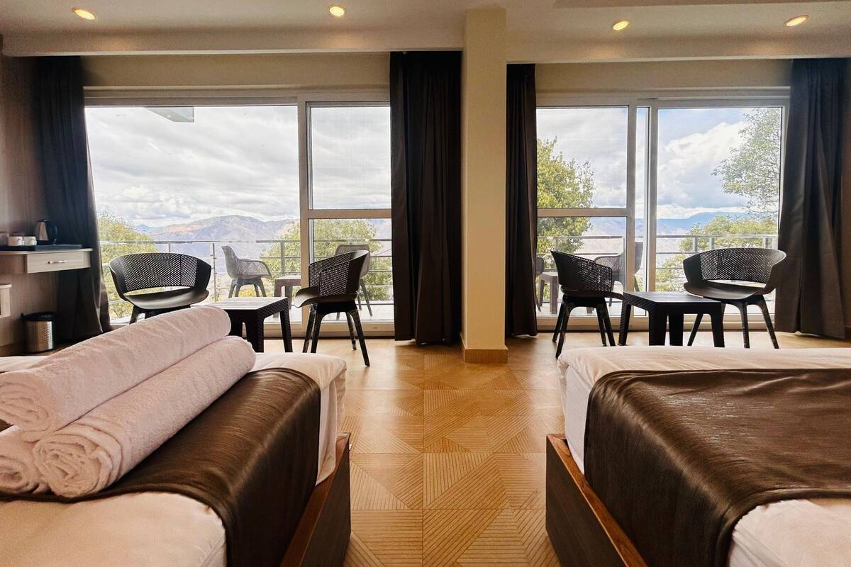 A hotel room with two beds, a desk, two chairs, and large windows with a view of mountains.