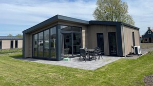 A modern building with large glass windows, a patio with outdoor furniture, and a tree nearby. - Modern Chalet With 2 Bathrooms (West-Graftdijk)