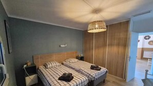 Une chambre avec deux lits, une tête de lit en bois, un luminaire suspendu et une porte donnant sur une autre pièce.