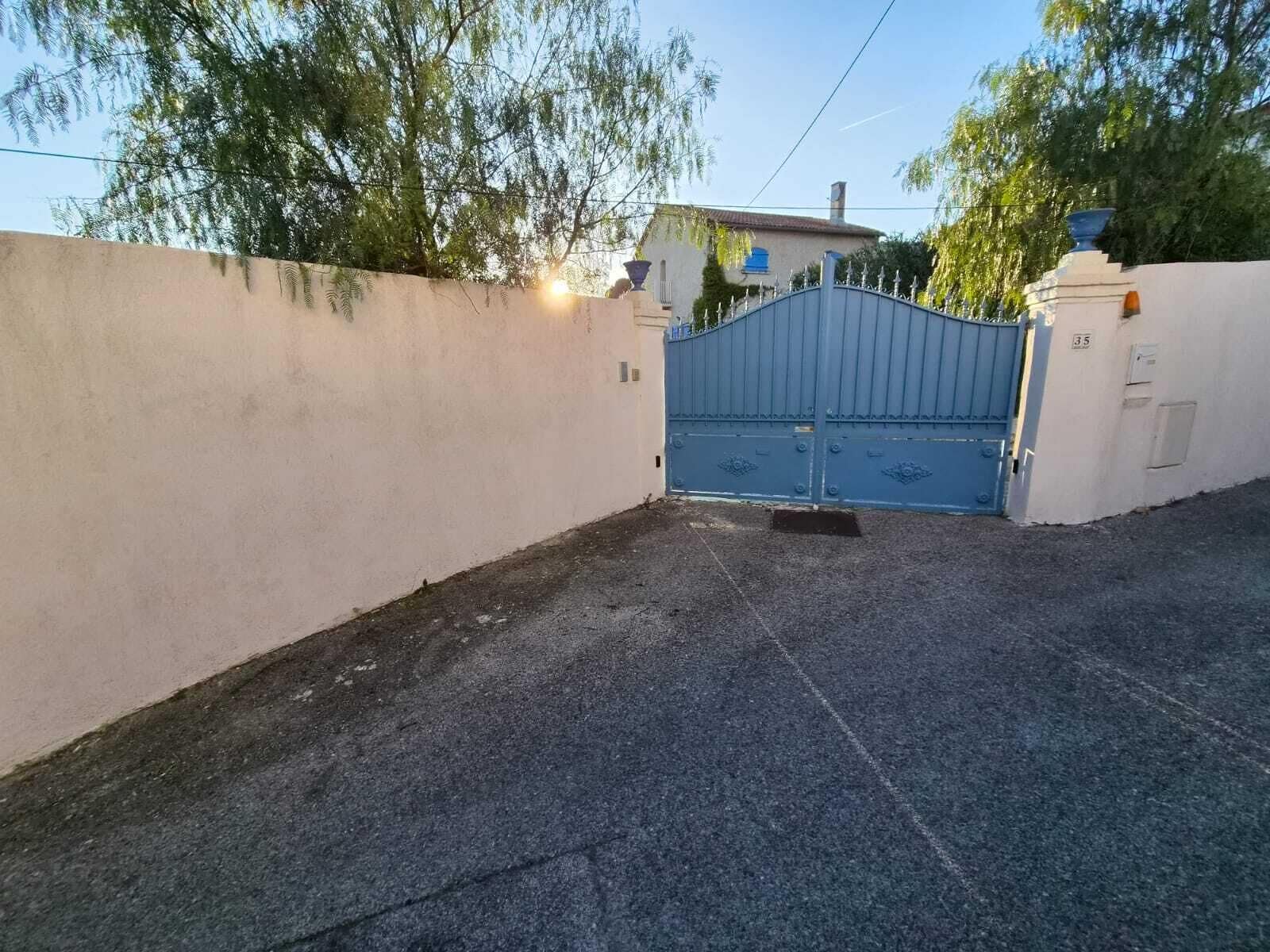 A gated driveway leading to a building with a white wall and a blue gate.