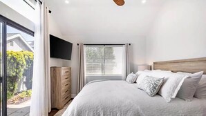Un dormitorio con una cama grande, un televisor montado en la pared, una ventana con persianas y vistas al exterior.