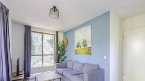 A living room with a grey sofa, a coffee table, a large window with curtains, a wall-mounted light fixture, and a painting of a landscape. - Lakeside Sauna Bungalow for 4 (Ewijk)