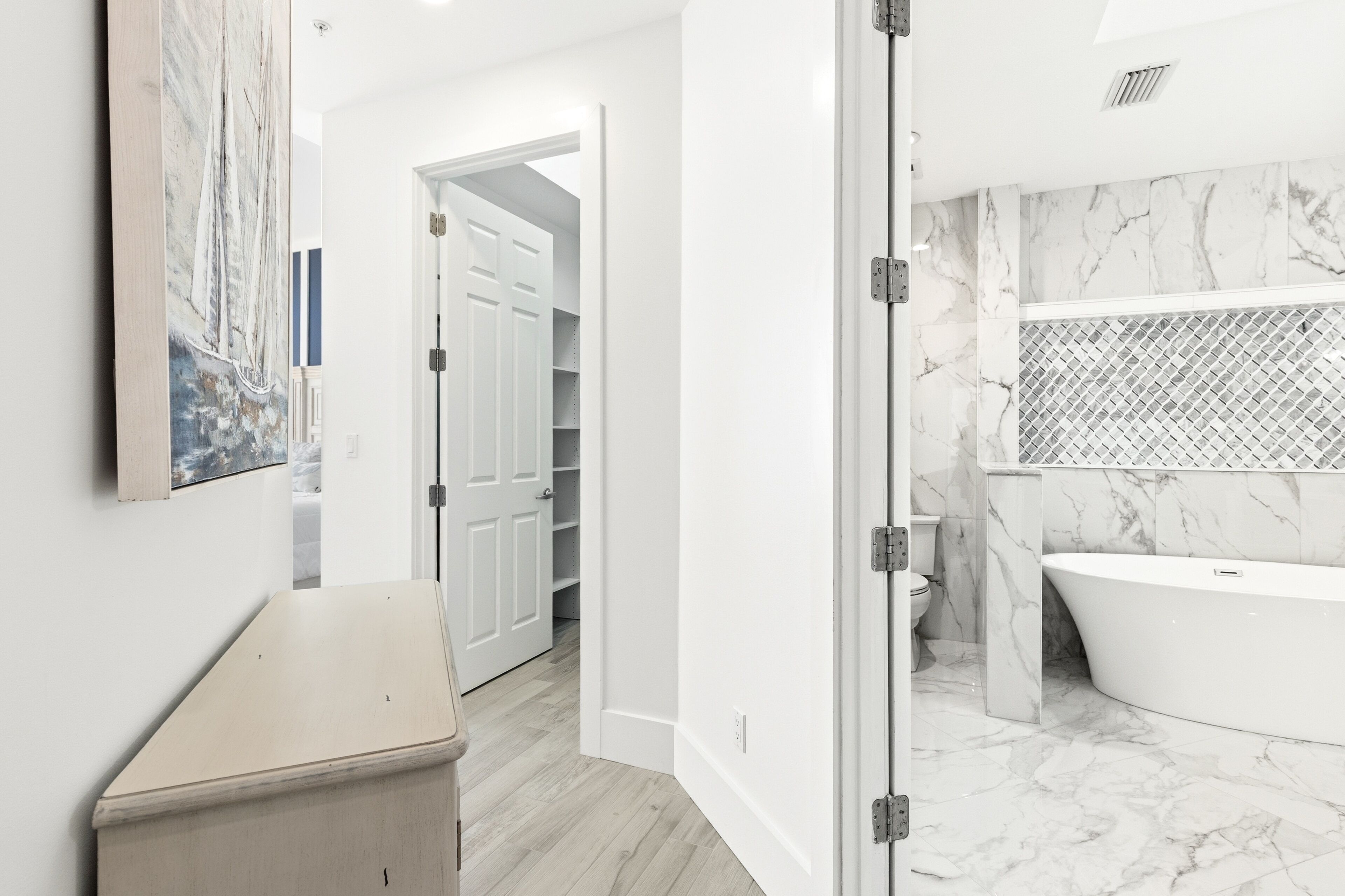 A modern bathroom with a freestanding bathtub, a marble backsplash, and a white door leading to a hallway with a white door and a closet.