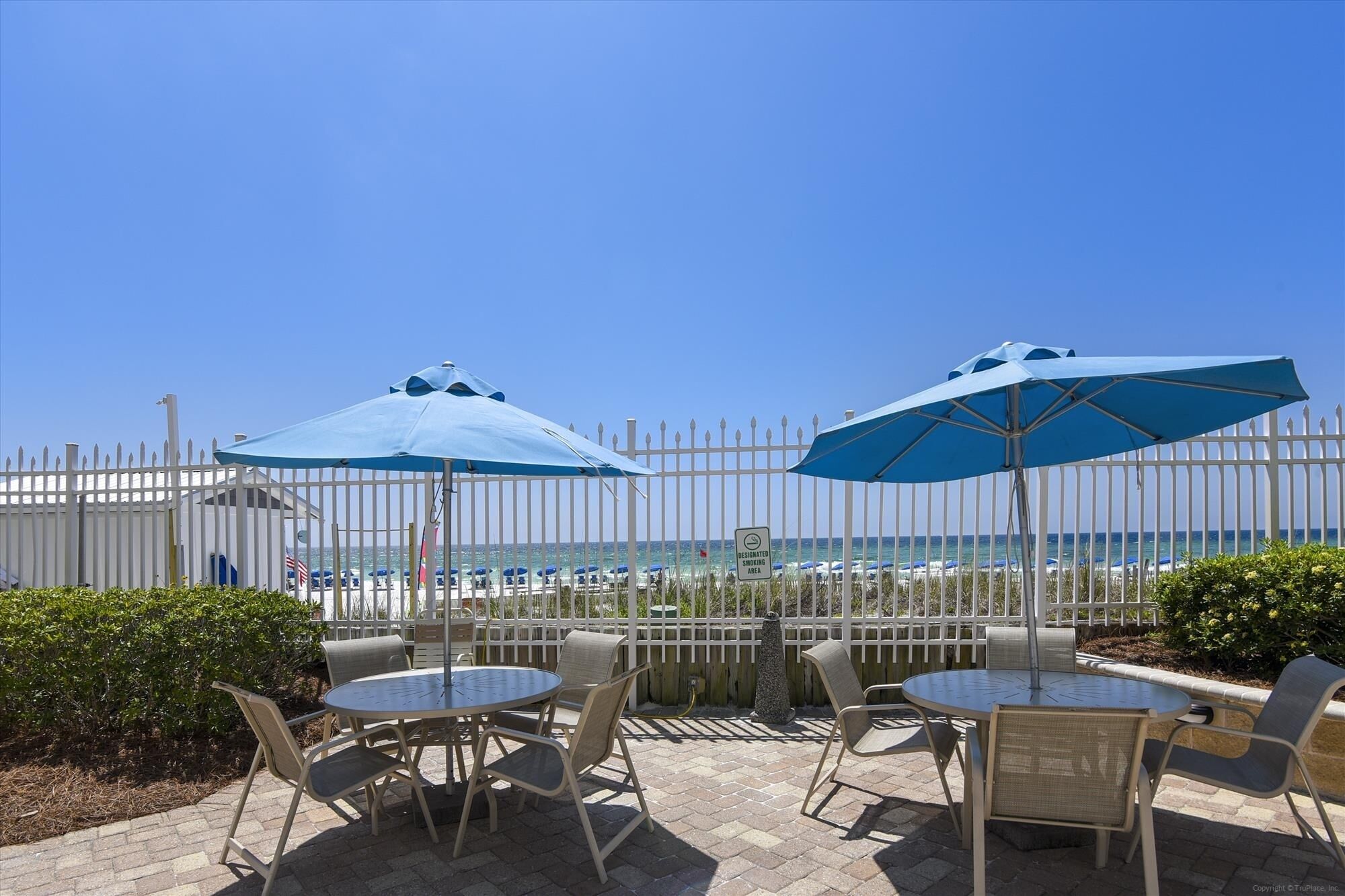 A patio with round tables and chairs, two blue umbrellas, and a view of the beach and ocean through a white fence.