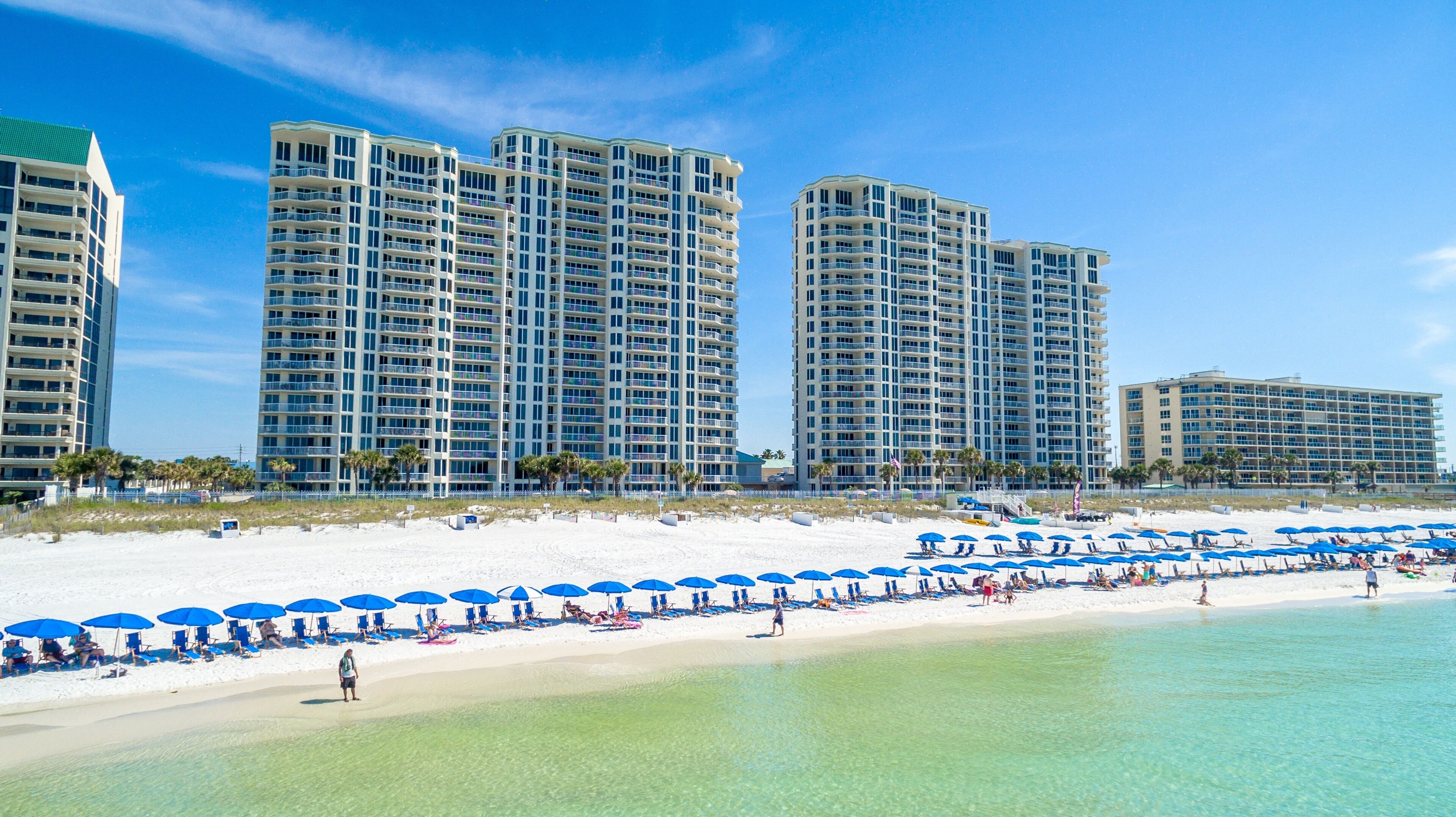 Een strandhotel met meerdere hoge gebouwen, een zandstrand met blauwe parasols en helder turquoise water.