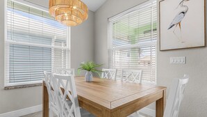 Un comedor con una mesa de madera, sillas blancas, una lámpara colgante y una obra de arte enmarcada en la pared.