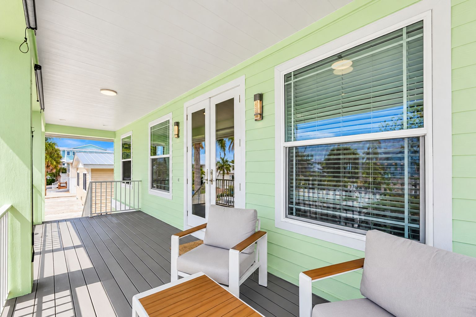 A covered porch with a wooden deck, light-colored walls, and large windows with blinds.