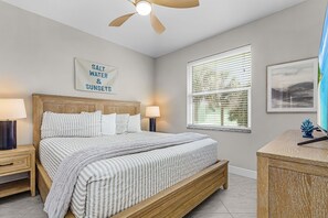 Een slaapkamer met een bed, nachtkastjes, een plafondventilator, een raam met zonwering en een lijst met een afbeelding aan de muur.