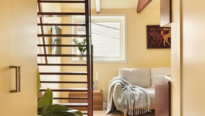 Un pasillo con un separador de listones de madera, un sofá, una ventana con persianas y un televisor instalado en la pared.