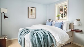 Un dormitorio con cama, una mesita de noche con lámpara, un cuadro enmarcado en la pared y una ventana con vistas a edificios.