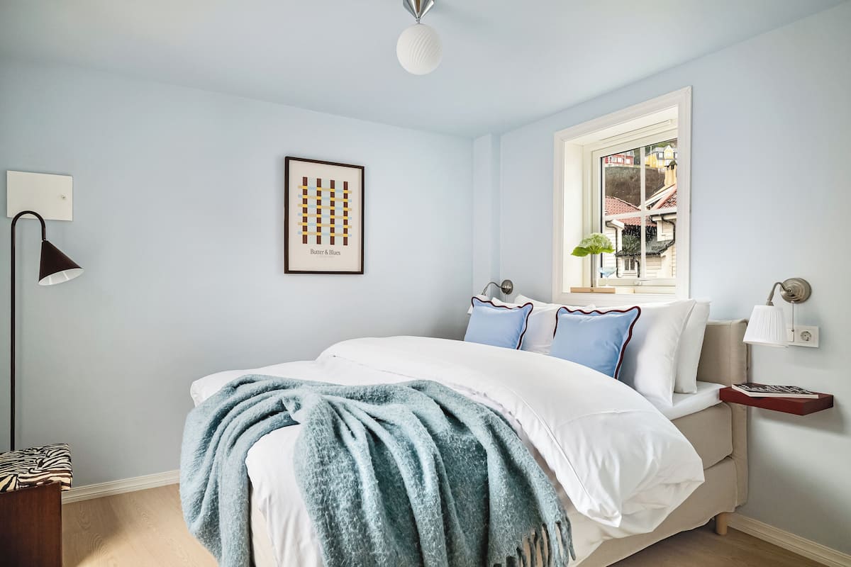 A bedroom with a bed, a bedside table with a lamp, a framed picture on the wall, and a window with a view of buildings.