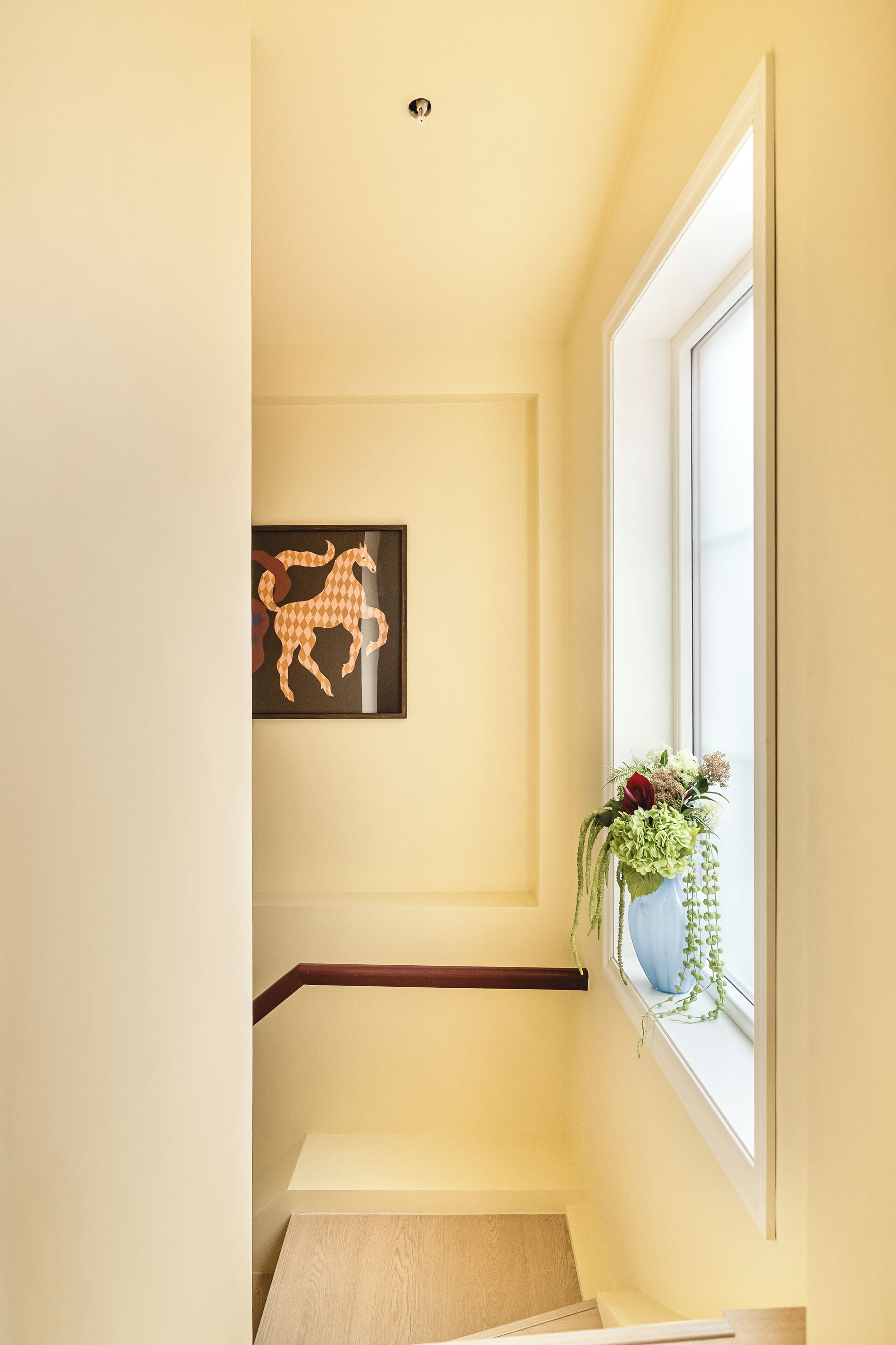 Una escalera con barandilla de madera, una obra de arte en el marco en la pared y una ventana con una planta.