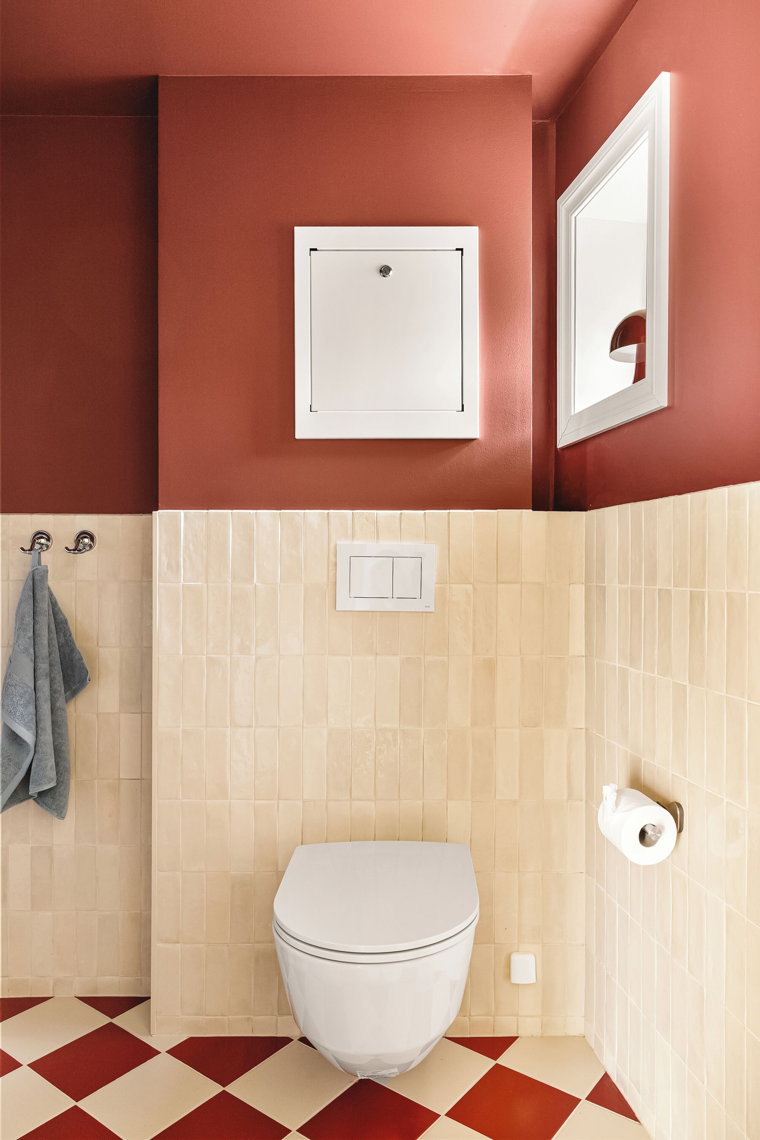 Une salle de bain avec des toilettes blanches, des murs carrelés de couleur beige, un sol à carreaux rouges et blancs, une serviette suspendue à un porte-serviettes et un porte-rouleau de papier toilette.