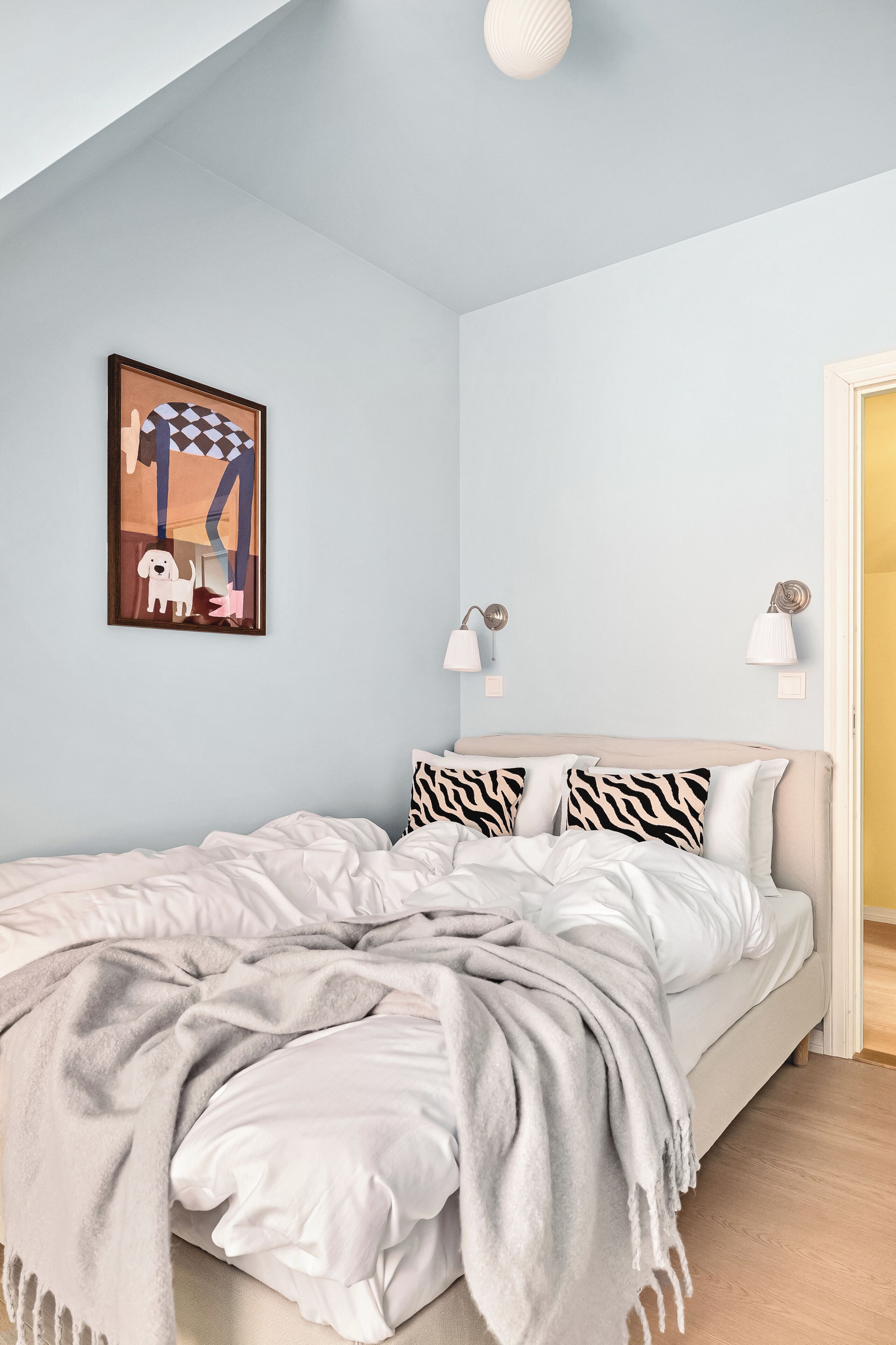 A bedroom with a bed, pillows, a blanket, a wall art piece, and two wall-mounted lamps.