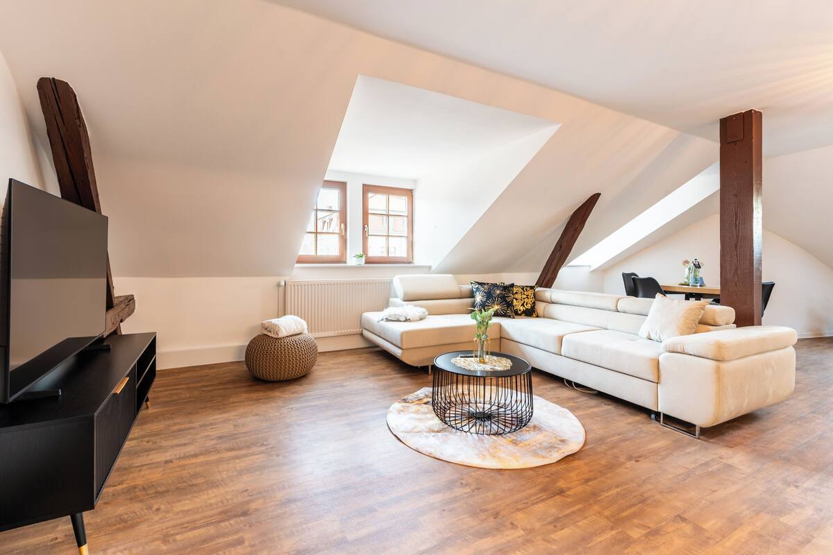A modern loft apartment with a high ceiling, a large window, a sofa, a coffee table, and a TV on a black stand.