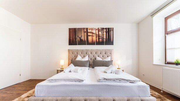 A double bed with a tufted headboard, two bedside tables with lamps, a large forest-themed artwork above the bed, and a window with blinds. - Vita Aurum Elbe-Apartments Neustadt  (Dresden)