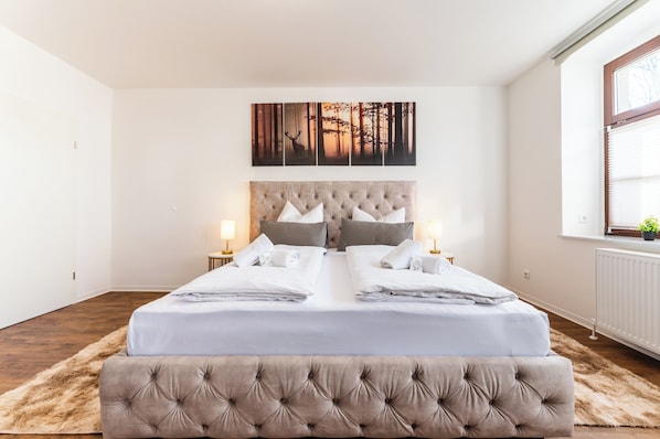 Ein Doppelbett mit einer gepolsterten Kopfteile, zwei Nachttische mit Lampen, ein großes Kunstwerk zum Thema Wald über dem Bett und ein Fenster mit Jalousien.