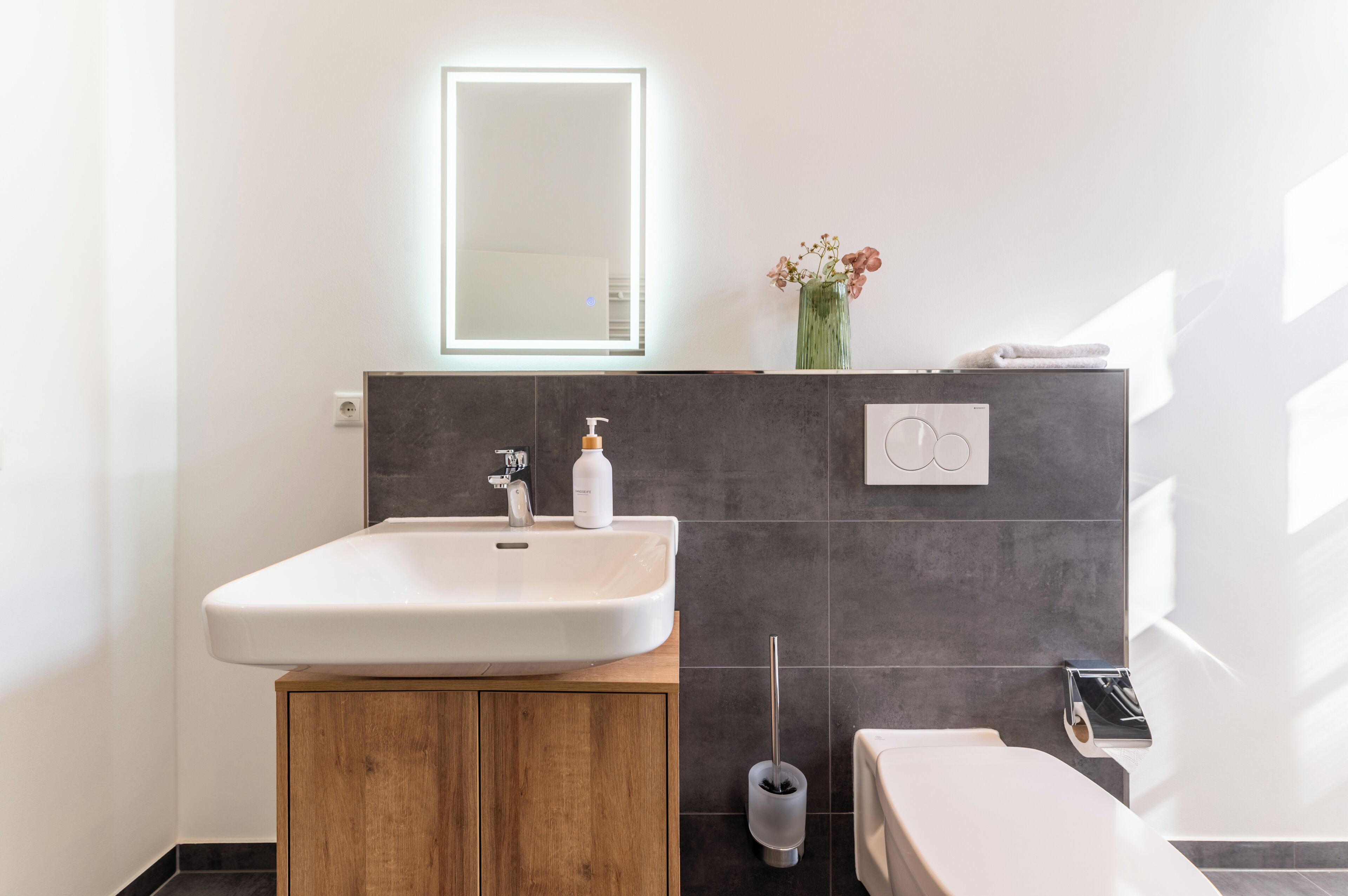 Une salle de bain avec un lavabo blanc, un meuble en bois et un miroir doté d’un éclairage intégré.
