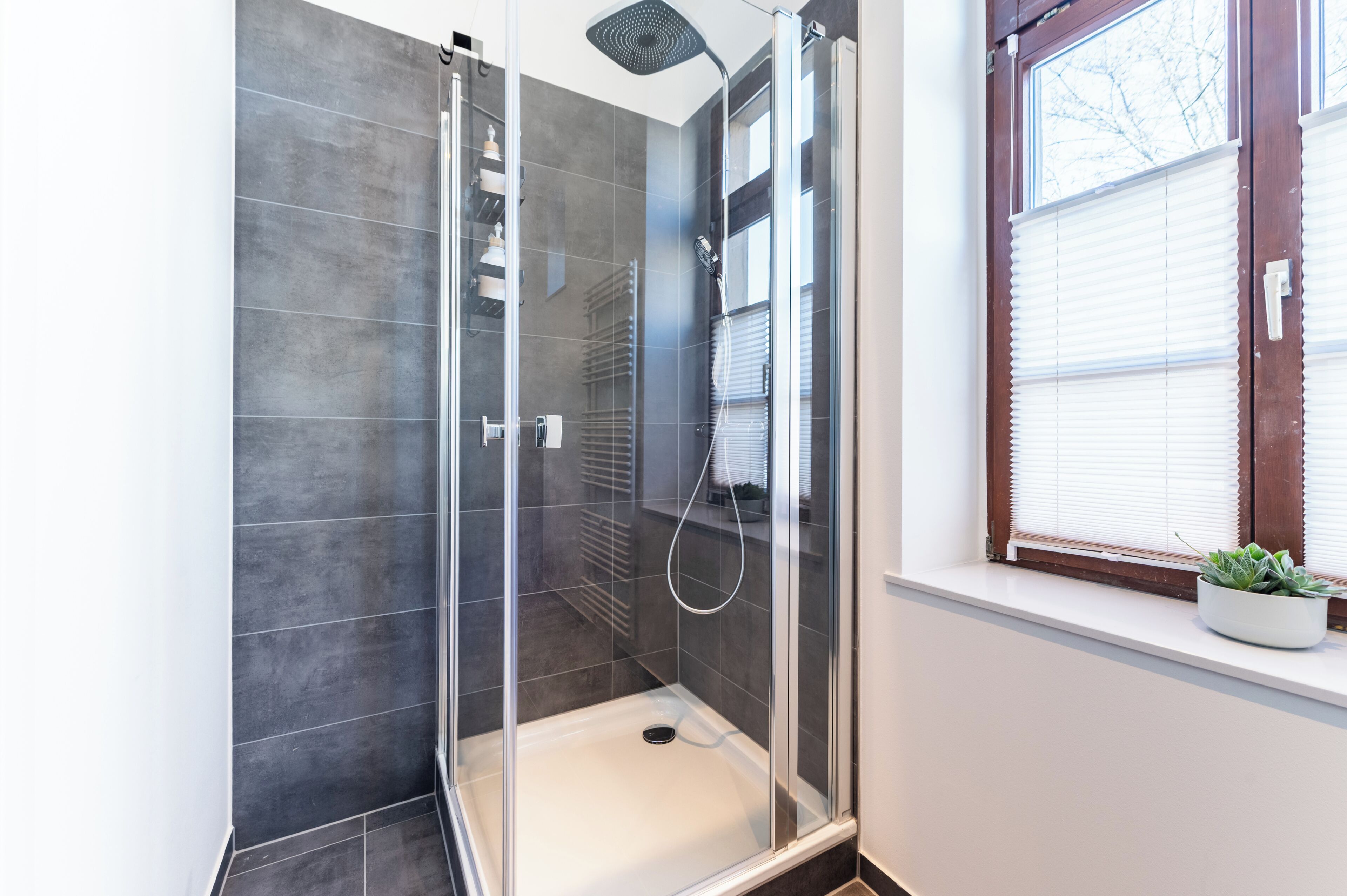 A glass-enclosed shower with a white floor, a window with blinds, and a potted plant on the windowsill.