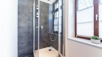 A glass-enclosed shower with a white floor, a window with blinds, and a potted plant on the windowsill.