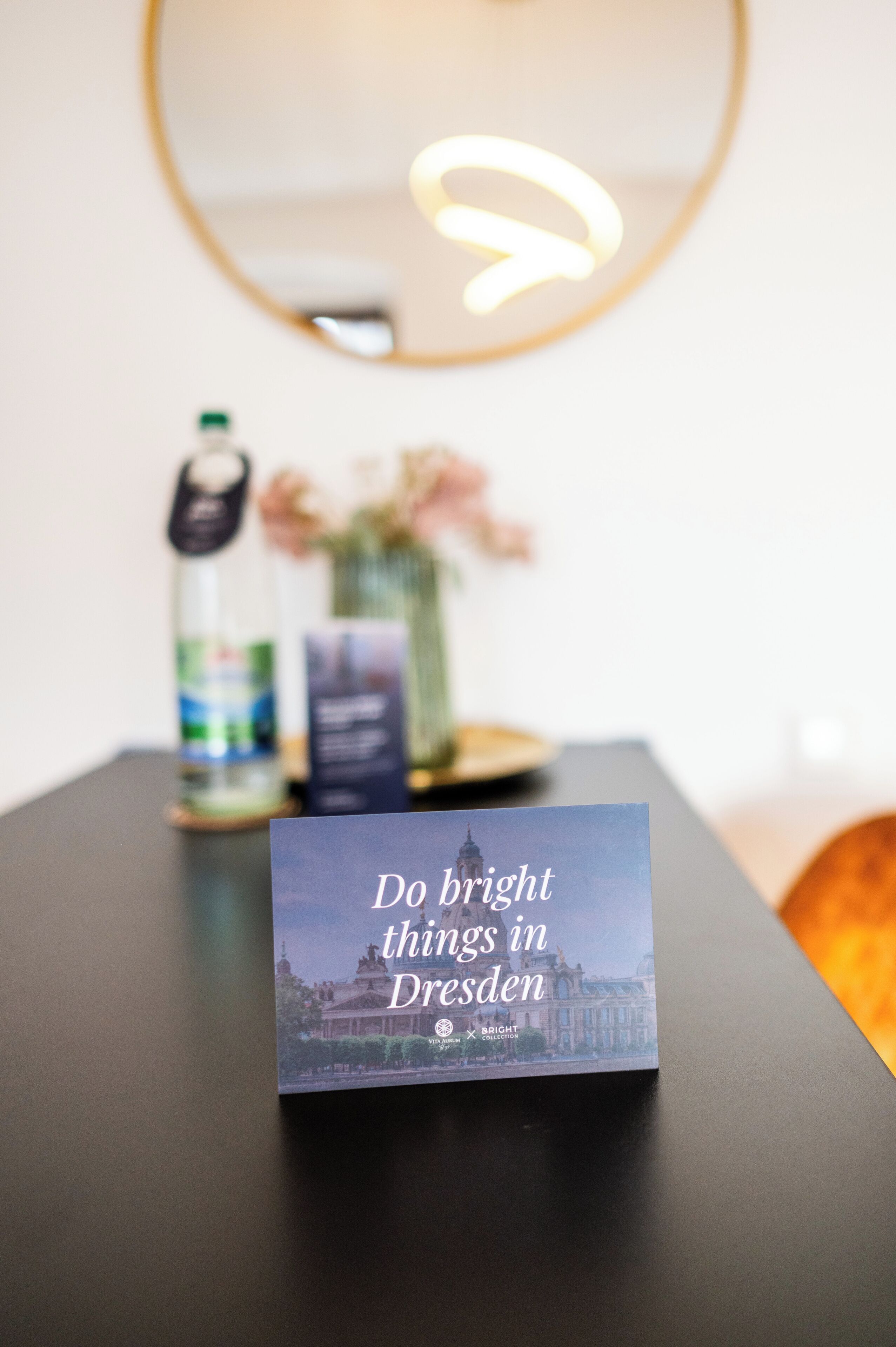 Un libro con el título 'Do bright things in Dresden' sobre una mesa con un espejo, una botella y un jarrón con flores.