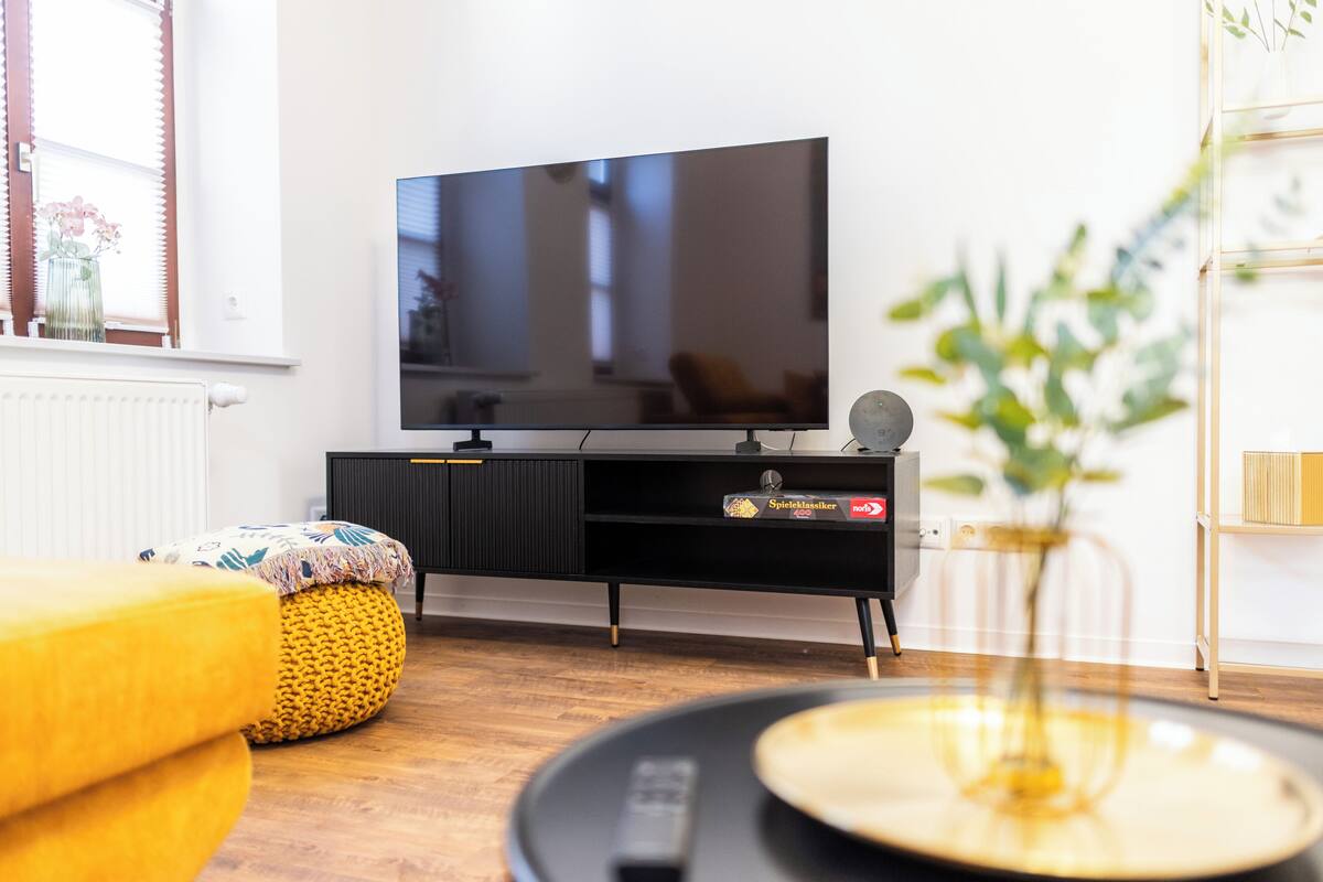 A modern living room with a flat-screen TV, a yellow ottoman, a black entertainment unit, and a plant.