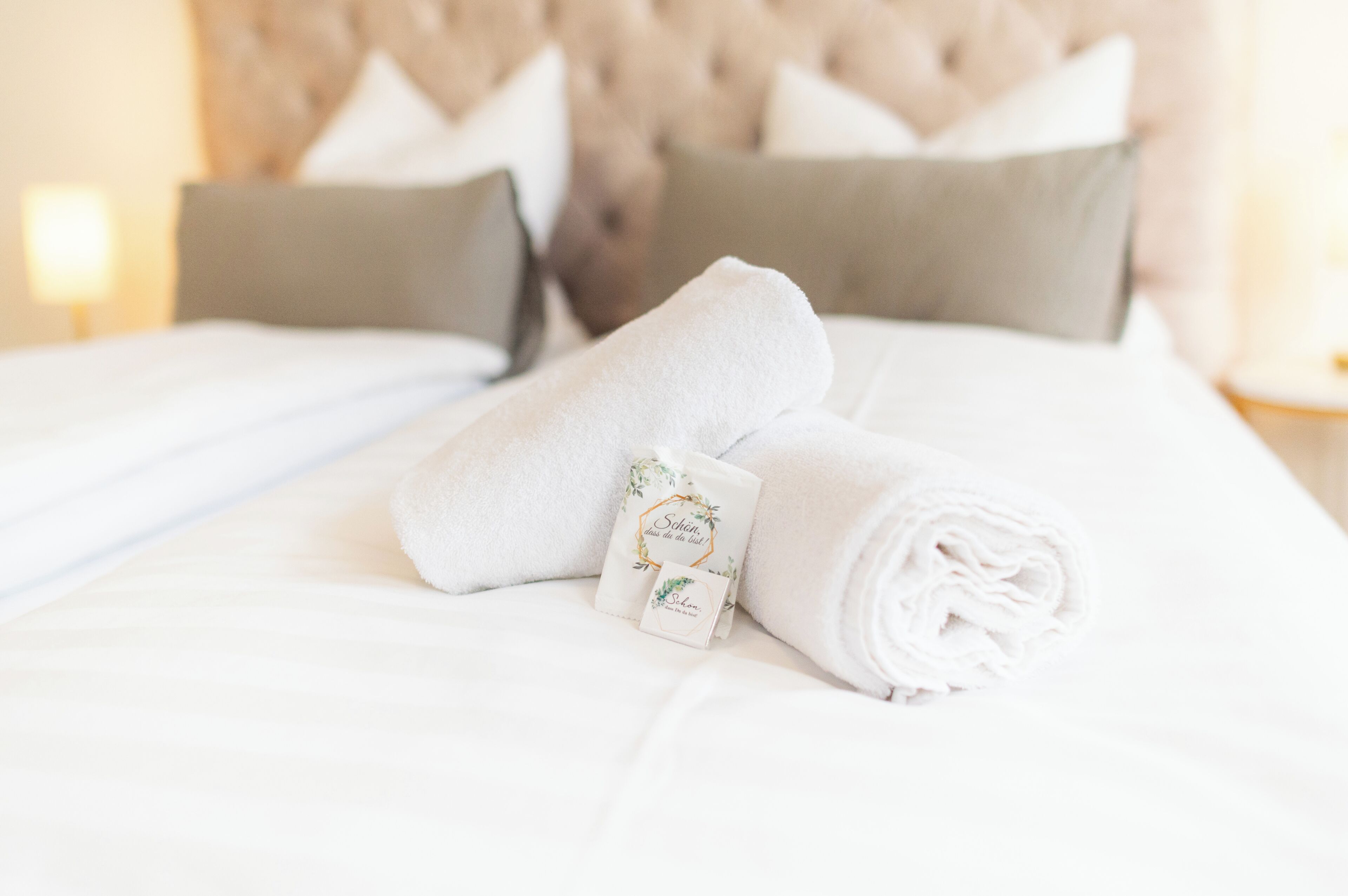 Een keurig opgemaakt bed met witte lakens, een opgerolde handdoek en een klein kaartje met een bloemendecor.