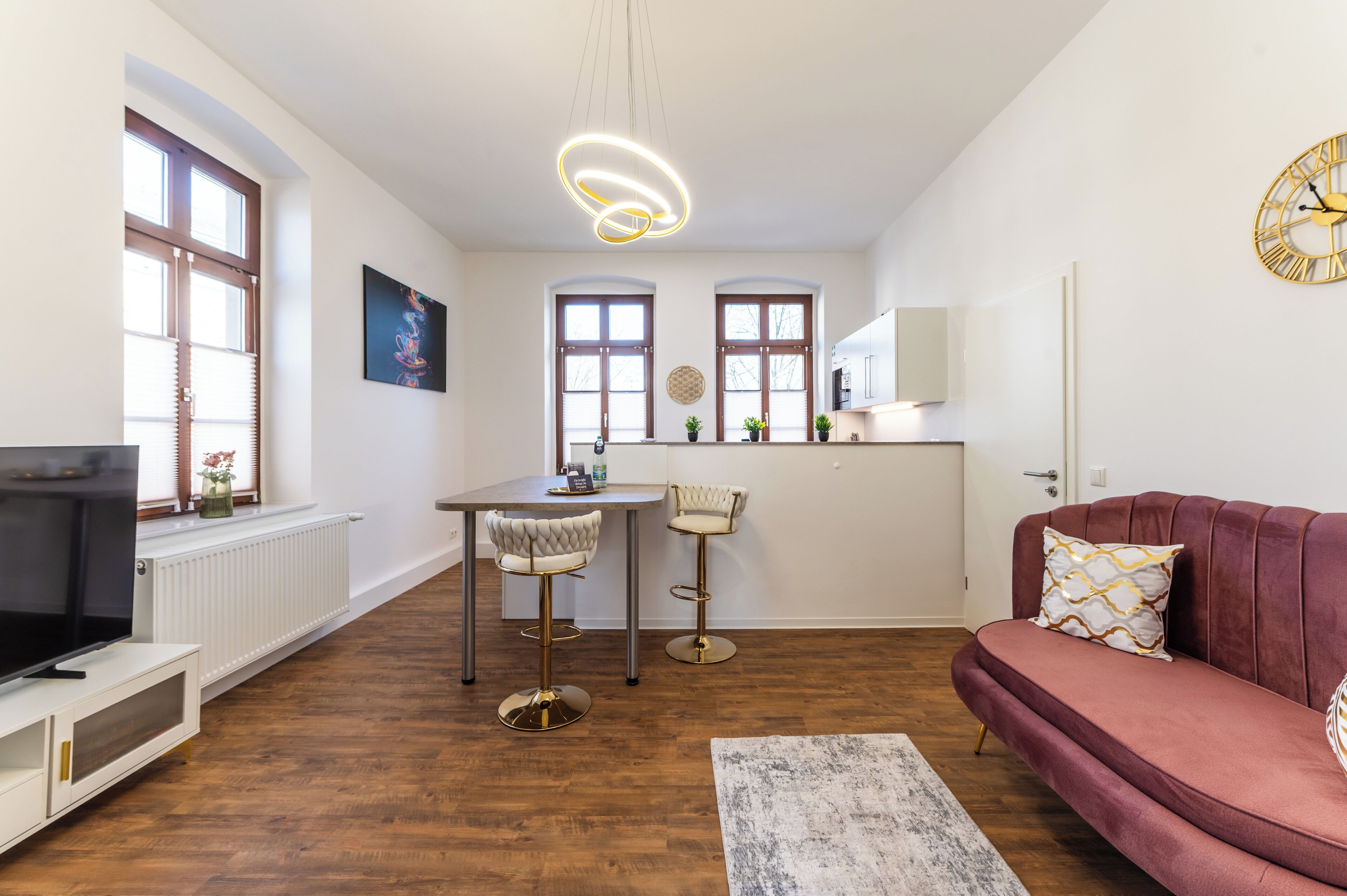 A modern living room with a sofa, a TV, a dining area with two chairs, a kitchen with a fridge, and a large window.