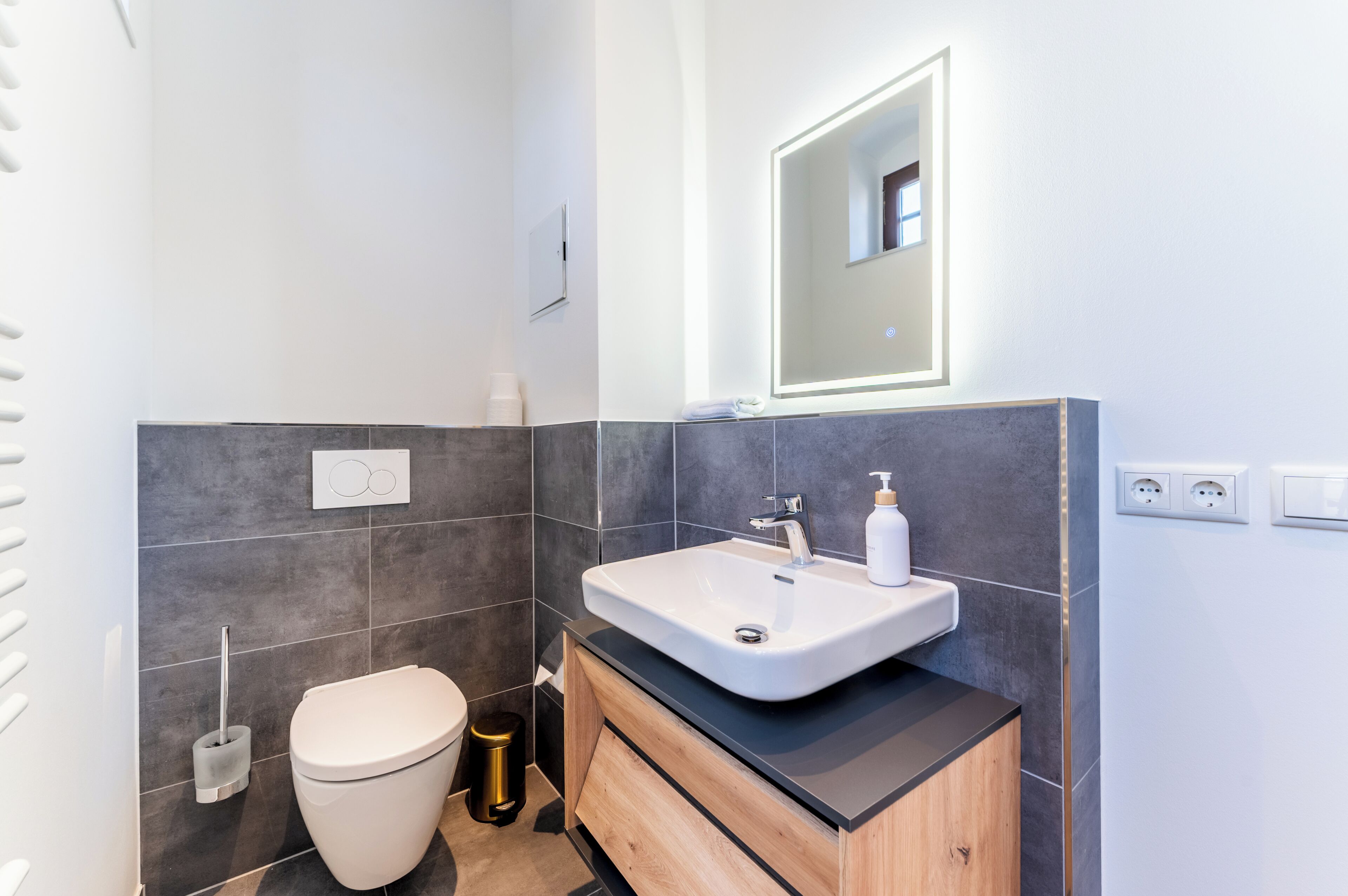 A modern bathroom with a white sink, a mirror, a toilet, and a trash bin.