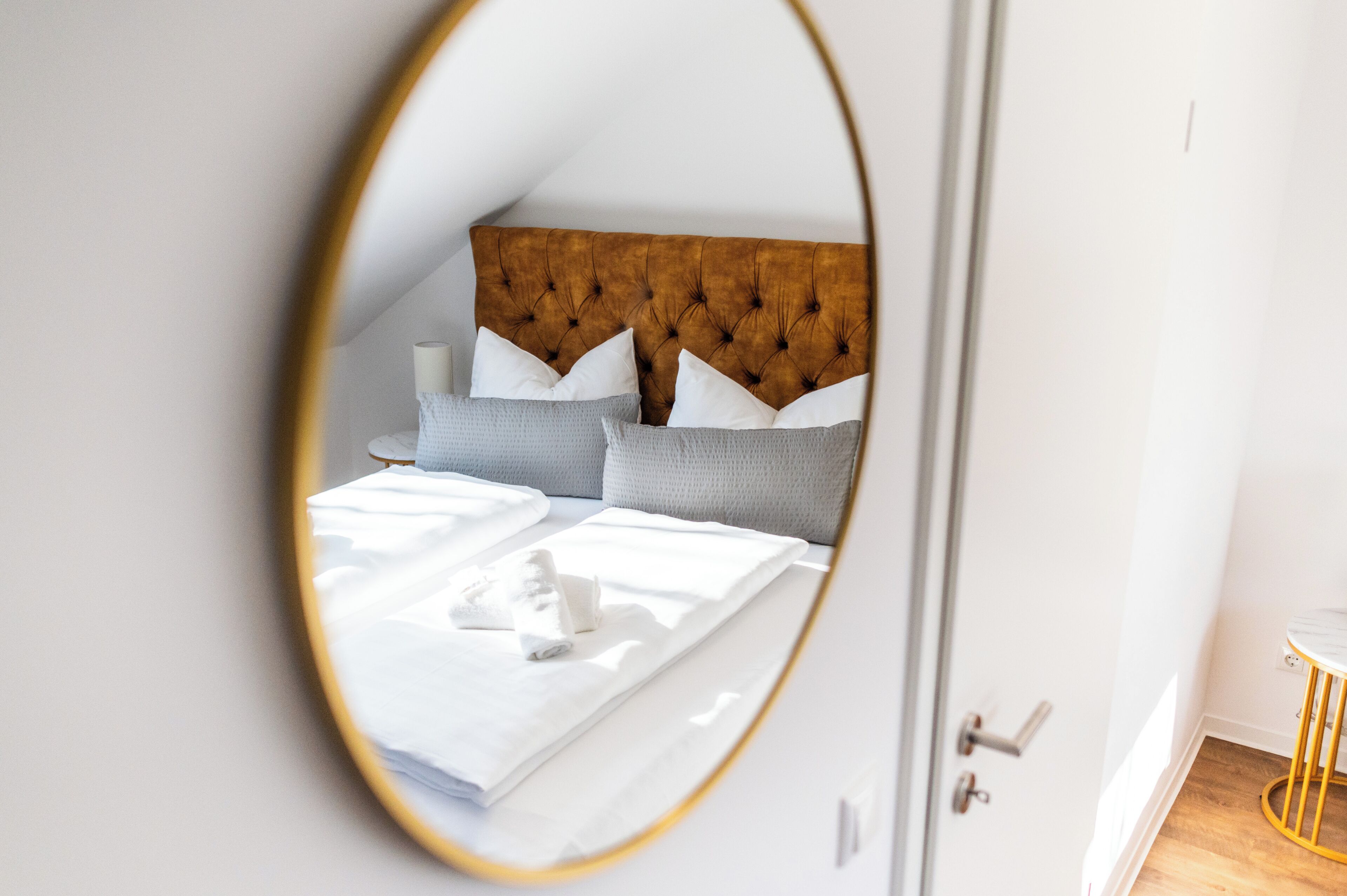 Een slaapkamer met een gestapeld hoofdbord, een bed met witte lakens en een ronde spiegel die de kamer weerkaatst.