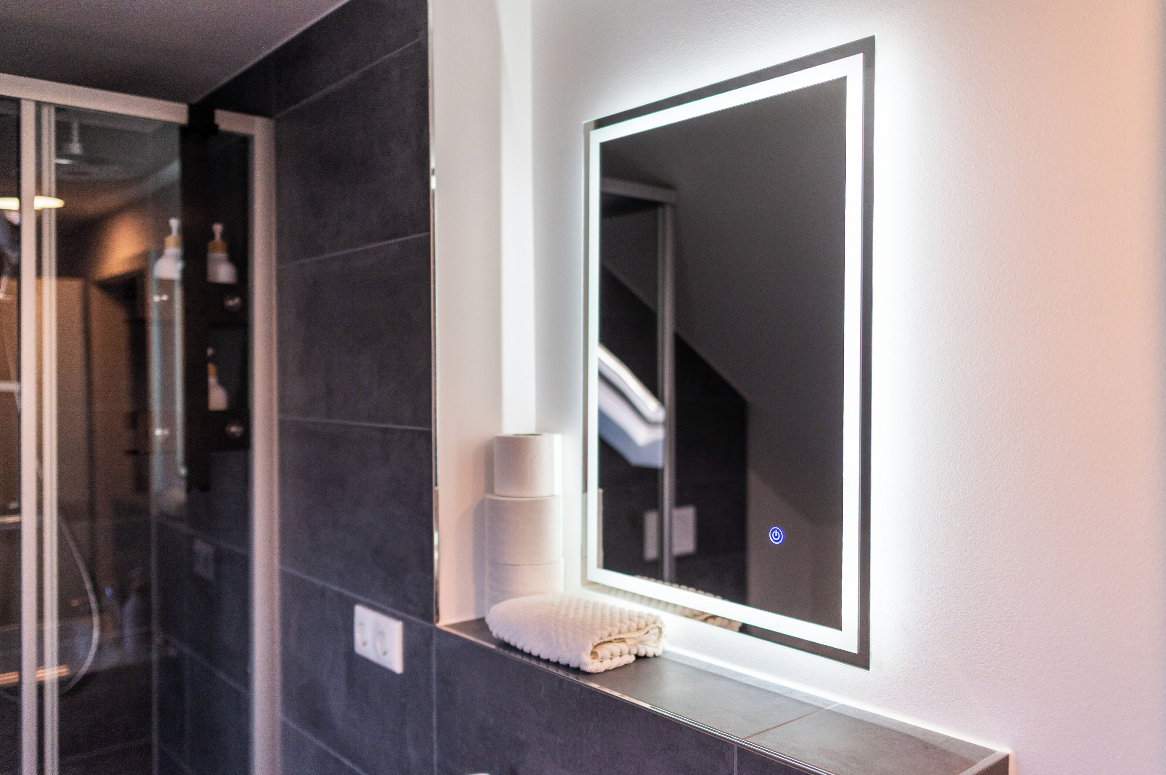 A bathroom with a large mirror, dark tiles, a towel on a shelf, and a glass shower enclosure.