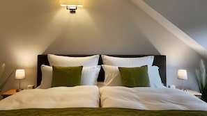 Un dormitorio con techo inclinado, una cama con ropa de cama blanca, dos almohadas verdes y lámparas de noche.