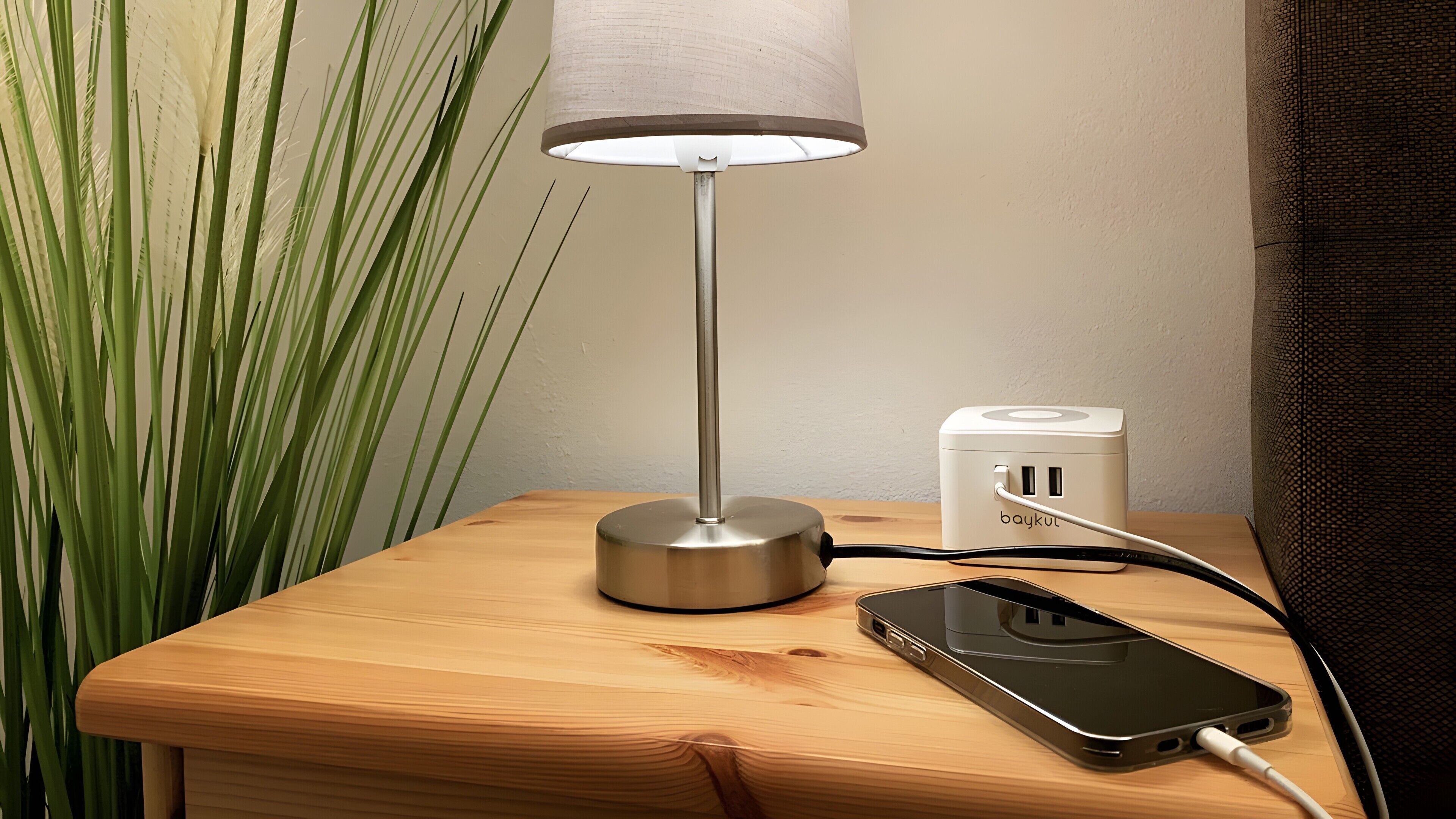 Una mesita de noche con una lámpara, un adaptador de corriente y un teléfono móvil cargándose.