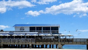 Un bâtiment situé en bord de mer, avec un toit bleu, une terrasse dotée de parasols et une jetée qui s’avance dans l’eau.