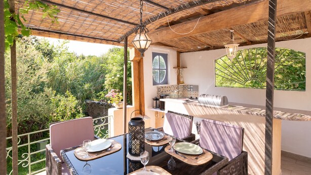 An outdoor dining area with wicker furniture, a table set with plates and glasses, a hanging lantern, and a view of greenery.