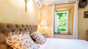A bedroom with a tufted headboard, floral pillows, a bedside lamp, and a window with a view of greenery.