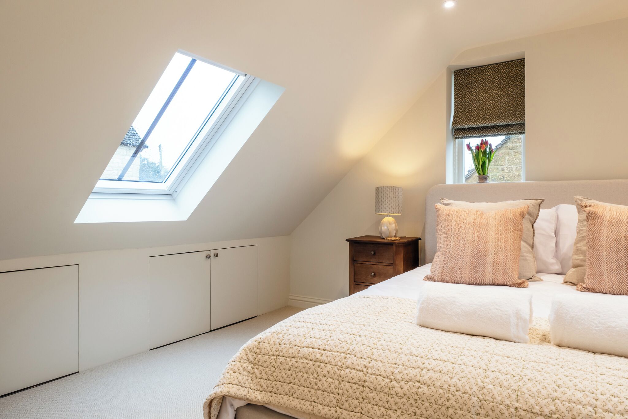 Un dormitorio con tragaluz, una mesita de noche con lámpara, una cama con almohadas y una ventana con vistas.