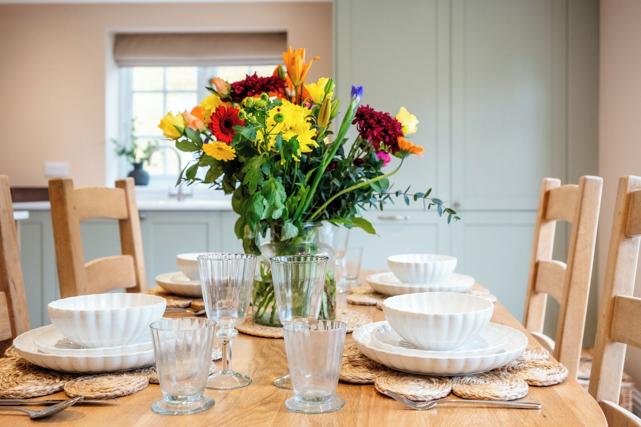 A dining table set with white plates, clear glassware, and a vase of colorful flowers.