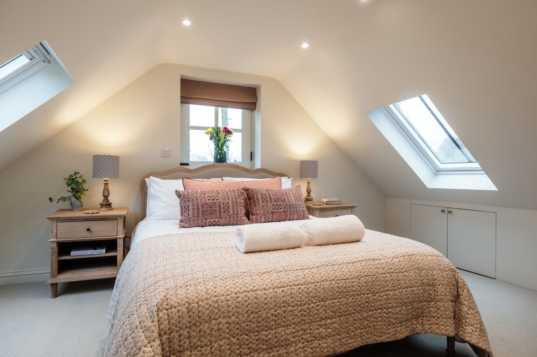 Camera da letto con soffitto spiovente, lucernari, un letto con coperta beige, due comodini con lampade e una finestra vista.