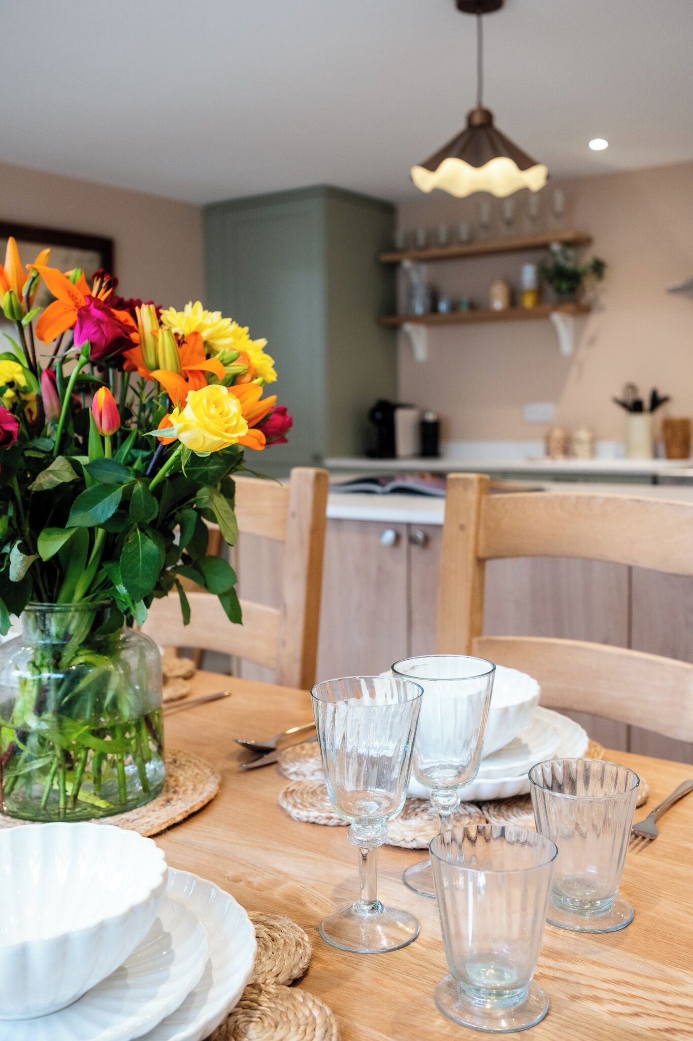 A dining table with a vase of flowers, clear glassware, and plates, set in a room with wooden furniture and a kitchen in the background.