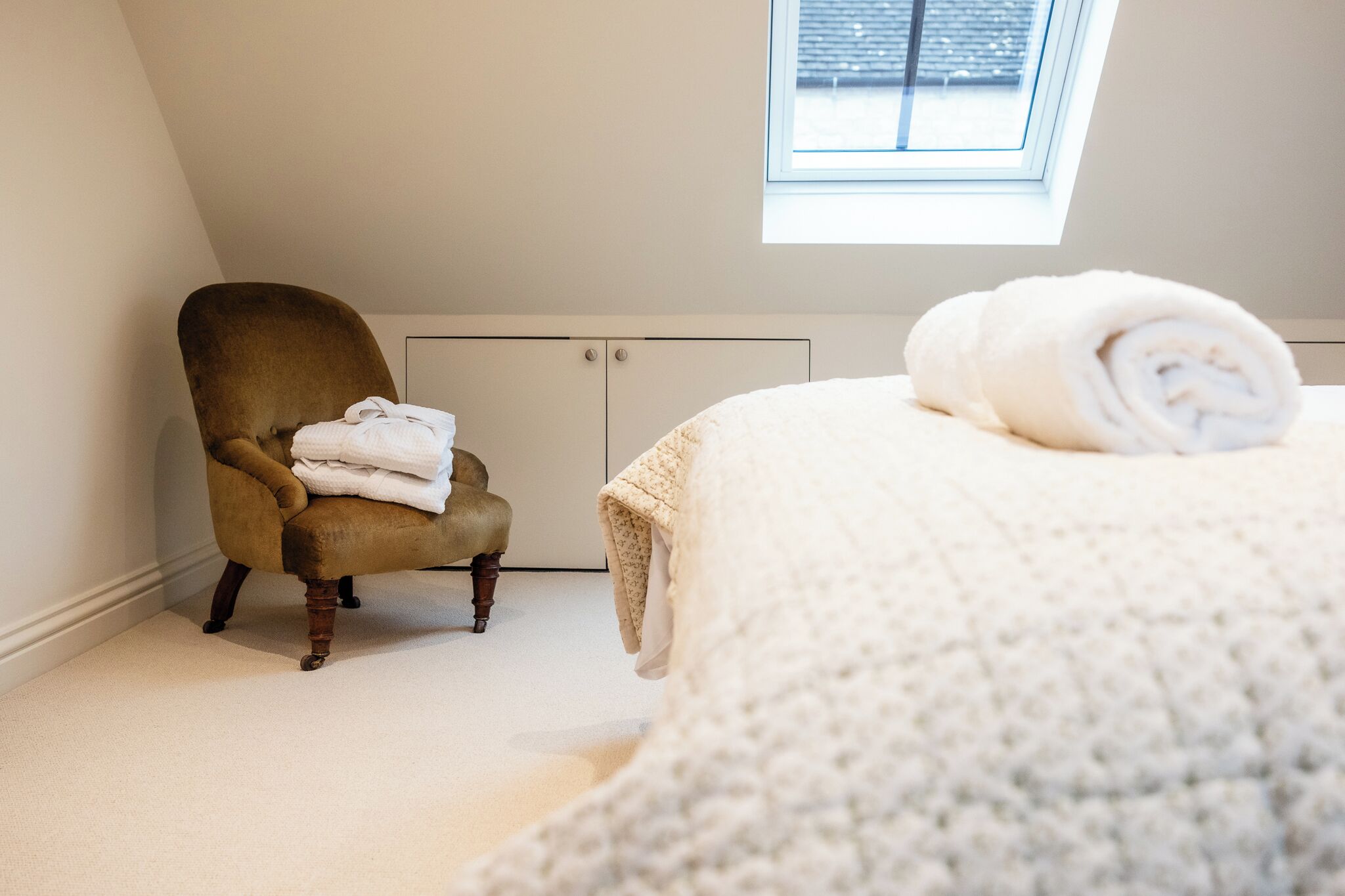 A bedroom with a high ceiling, a window, a radiator, a chair with towels, and a bed with a blanket.