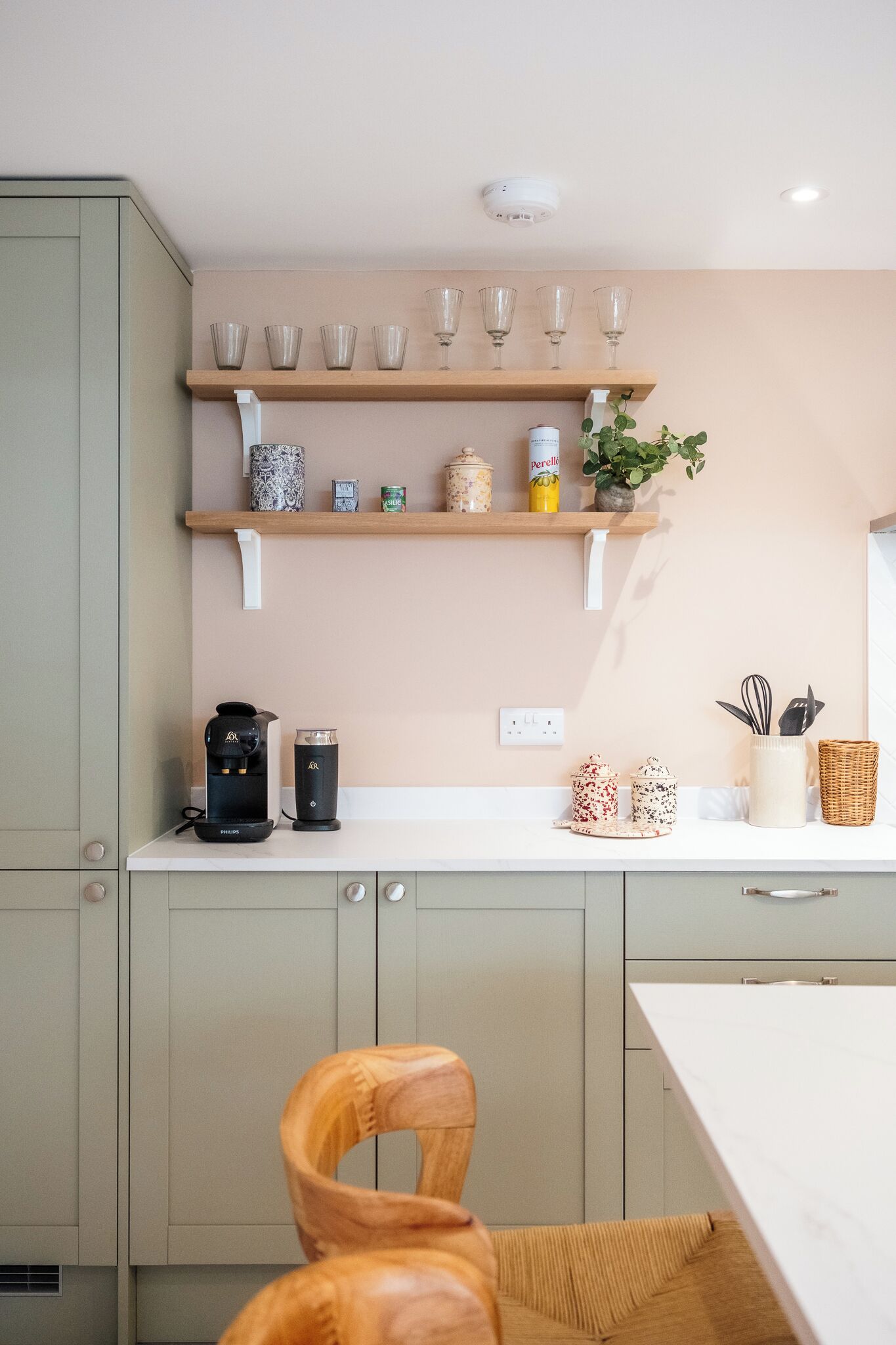 Una cucina con un piano di lavoro bianco, due macchine da caffè, uno scaffale con bicchieri e una sedia in vimini.