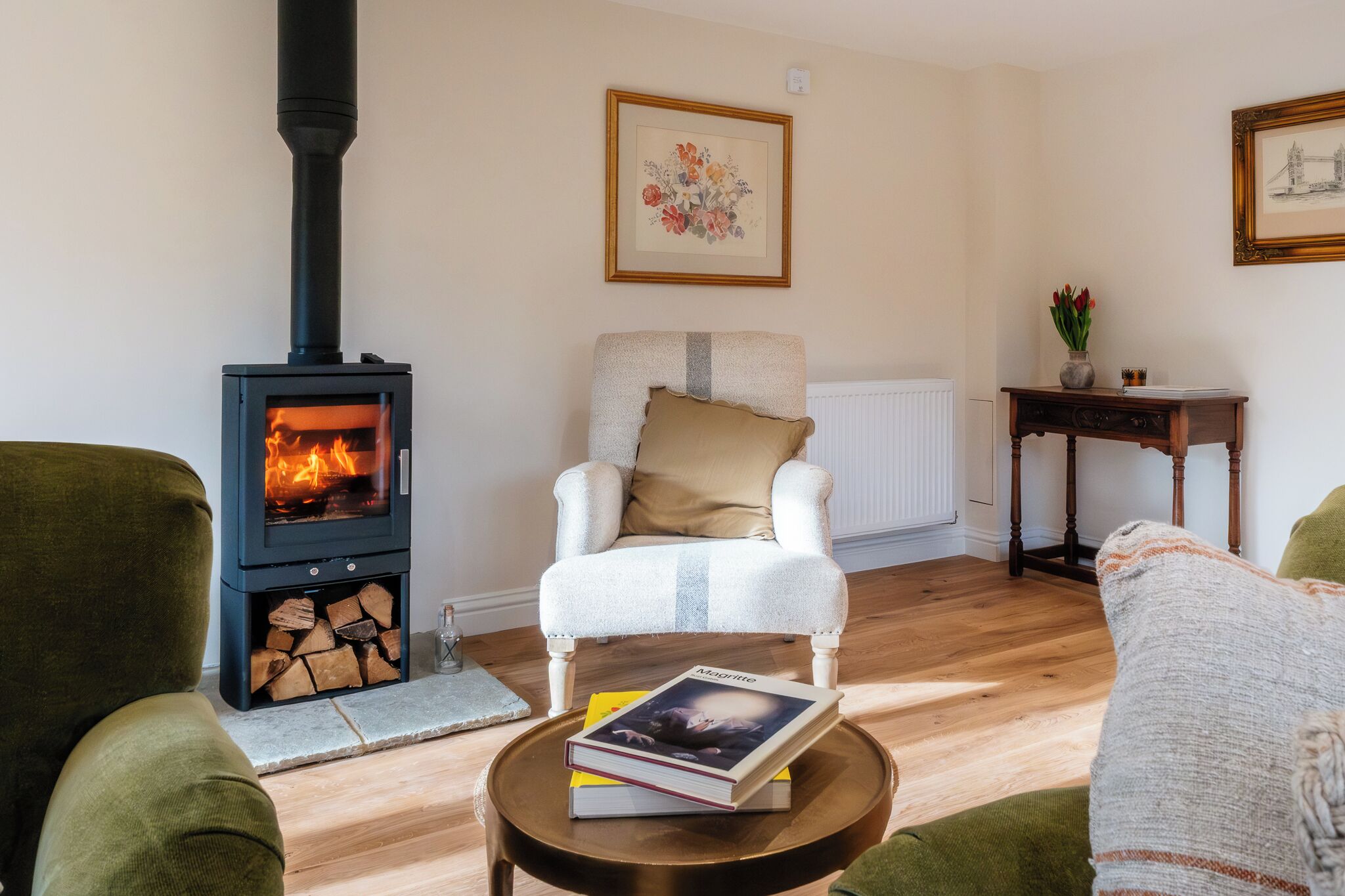 A cozy living room with a fireplace, a comfortable armchair, a small table with a book, and a framed picture on the wall.